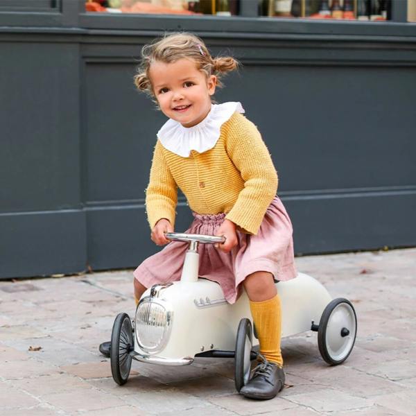 A young girl with pigtails wearing a mustard yellow cardigan and pink skirt, smiling while riding a vintage cream-coloured metal ride-on roadster car on a stone pavement path.