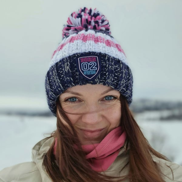 Woman in snowy tree lined glade in Northern Finland is wearing a navy, white and pale pink striped chunky knit Super Bobble hat.  The hat also has a deep ribbed headband and contains reflective yarns and a luxury sherpa fleece lining.