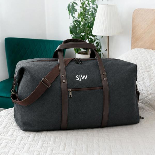 A personalised dark grey washed canvas weekend bag with brown handles and strap, embroidered with white initials SJW, resting on a white textured bedcover.