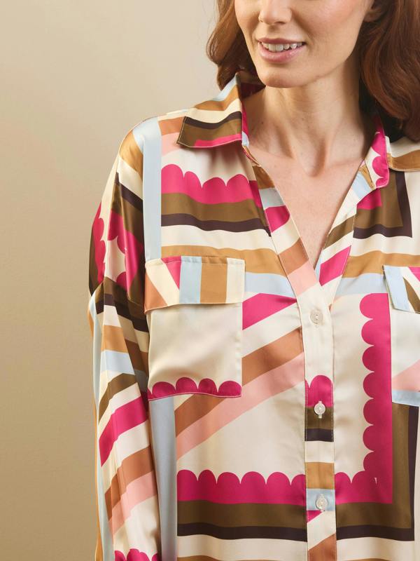 A close-up of a woman wearing a long-sleeve button-up pyjama shirt with a retro striped pattern in pink, brown, beige, and light blue. The shirt features scalloped edges along the pocket flaps and collar.