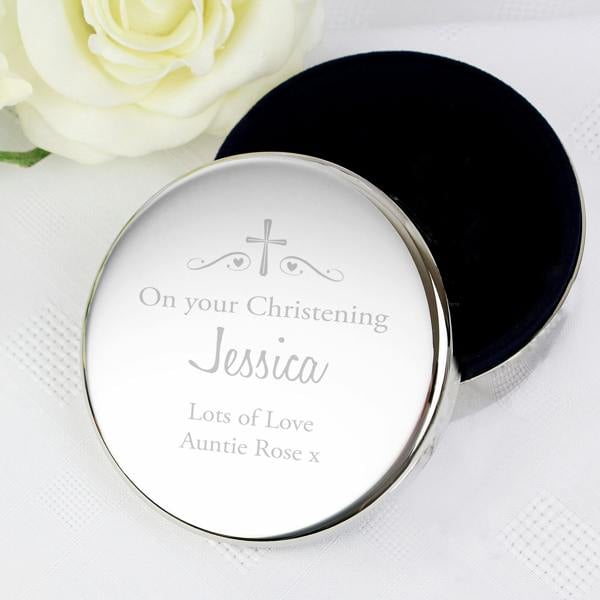 A round nickel-plated christening trinket box by Abode Home & Gifts, resting slightly open on a white linen tablecloth next to a white rose. The lid is custom engraved with a cross, heart scrolls, and the text "On your Christening Jessica Lots of Love Auntie Rose x".