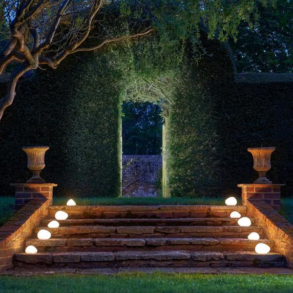 Multiple frosted glass solar pebble lights illuminating symmetric brick and stone garden steps leading toward a large hedge archway at night.