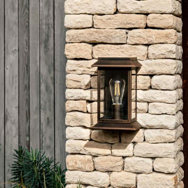 Antique bronze steel lantern wall light with a classic glass frame and filament bulb, mounted on a light-coloured stone wall next to grey wooden panelling.