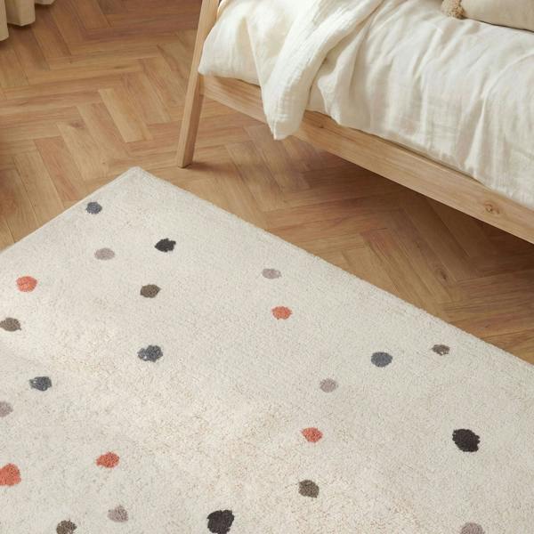 Beige rug with multicolored dots on a wooden floor next to a bed.