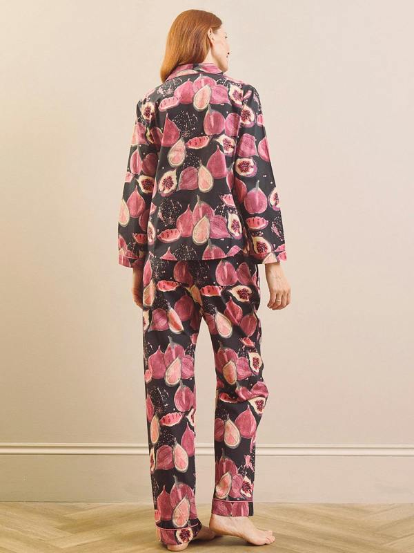 A woman stands with her back to the camera wearing a matching pyjama set with a pink and red fig pattern on a black background, set against a neutral beige wall and light wood floor.