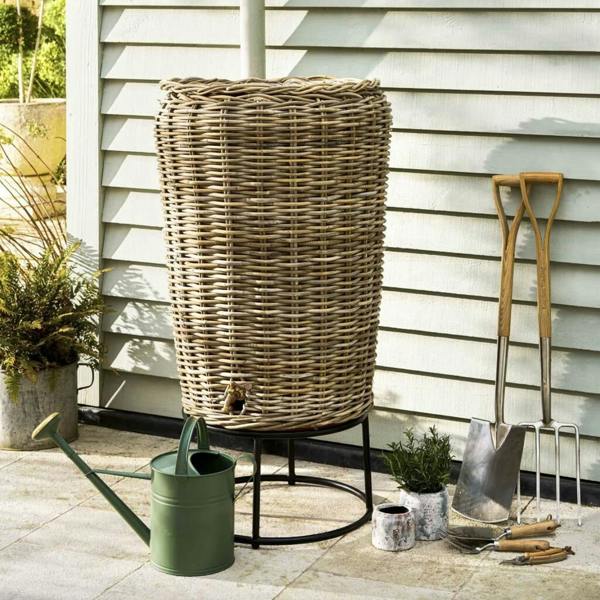 A tall, woven grey rattan water butt sitting on a black metal stand next to a green metal watering can and wooden garden tools against a cream shiplap wall.