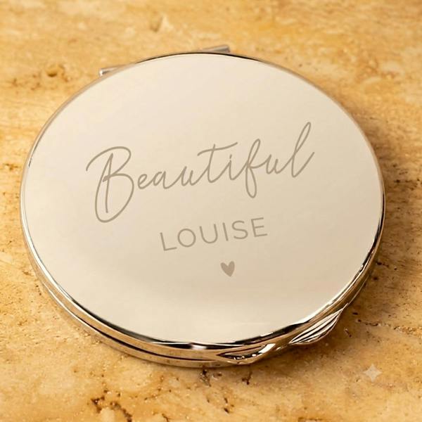 A polished silver personalised compact mirror engraved with 'Beautiful Louise' and a heart motif, resting on a warm beige stone surface.