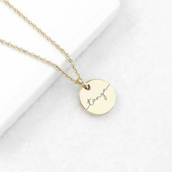 An 18ct gold plated disc necklace engraved with the name 'Tanya' resting diagonally across a clean white marble slab.