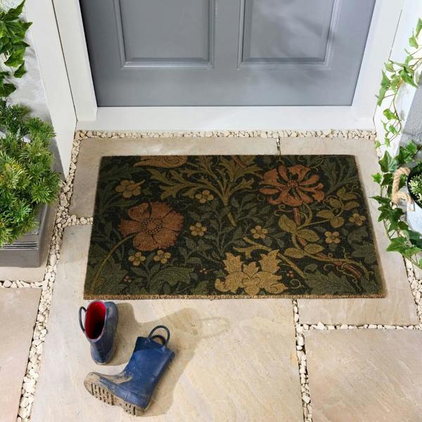 Decorative doormat with floral pattern in front of a gray door, with boots and a plant on a stone path.