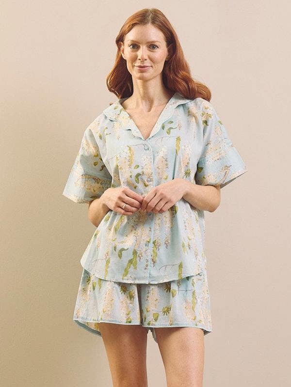 A woman modelling a light blue short-sleeve pyjama set with a light blue and yellow floral print, consisting of a button-up top and matching shorts, against a solid beige background.