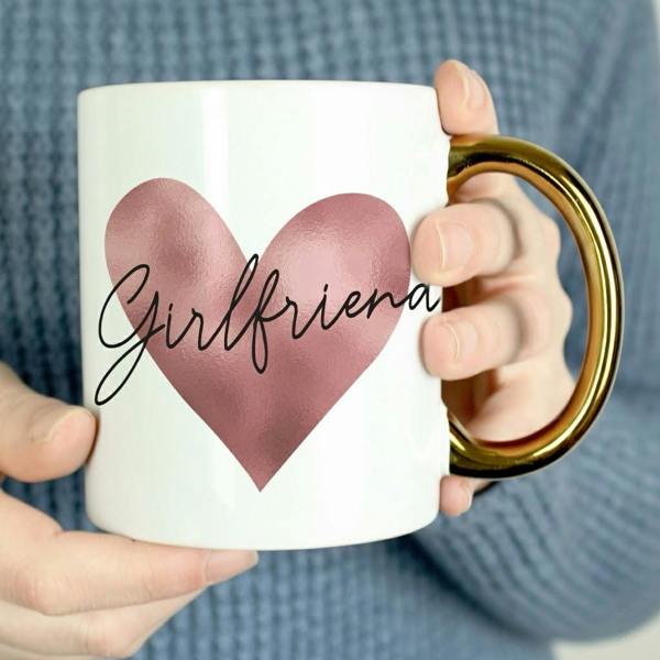 A close-up of a person holding a white ceramic mug with a luxury gold handle and a pink metallic heart printed with the name Girlfriend in cursive script.
