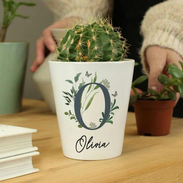 Custom white ceramic plant pot featuring a botanical letter O design and the name Olivia holding a green cactus on a wooden surface.