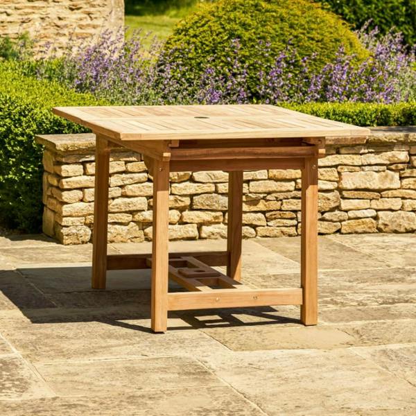 Full side profile of the rectangular solid teak extending dining table standing on a stone patio.
