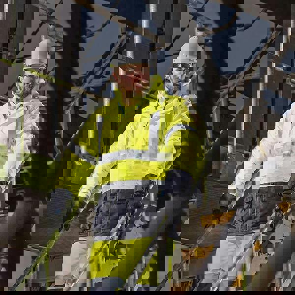 Scruffs High Visibility Mens Hi-Viz Waterproof Zip Up Jacket Coat, a man wearing a bright yellow high visibility waterproof jacket with reflective stripes, hard hat, and gloves at an industrial site.