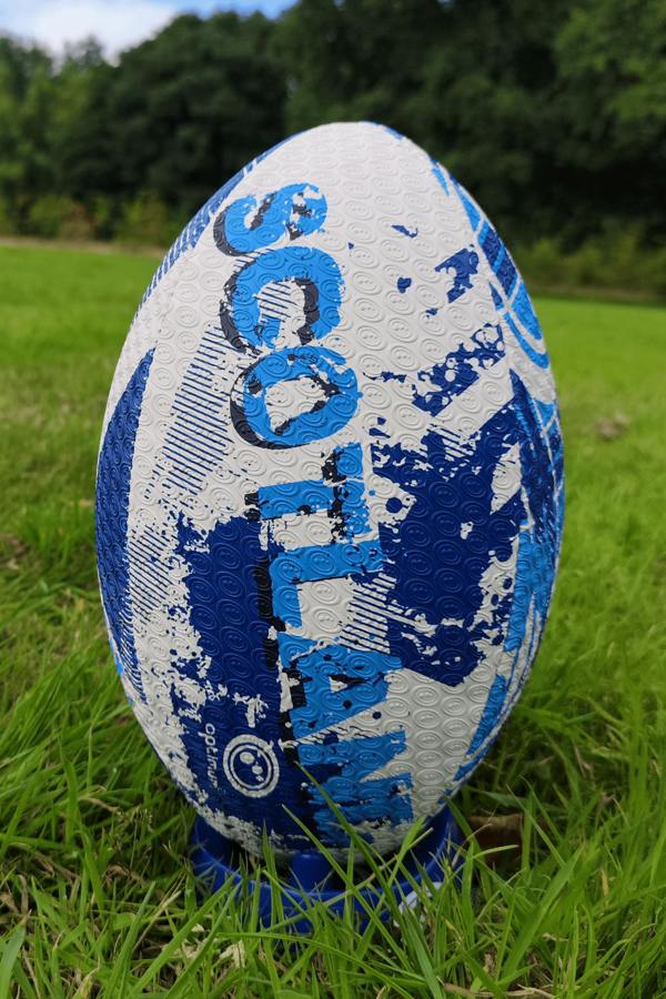 White and blue Optimum Scotland rugby ball upright on a kicking tee in grass with trees behind - Optimum