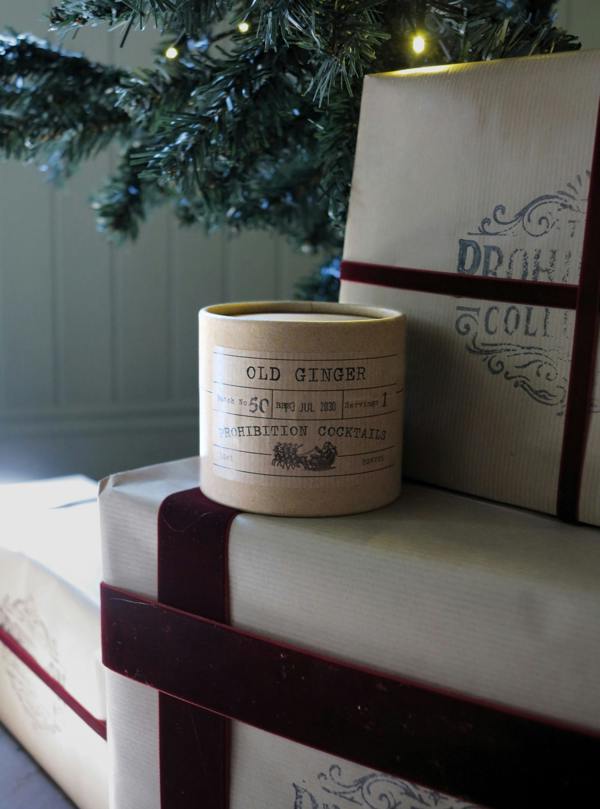 A cylindrical candle jar labeled 'OLD GINGER' sits on top of wrapped gift boxes with dark red ribbon, with a Christmas tree branch and warm string lights in the background.