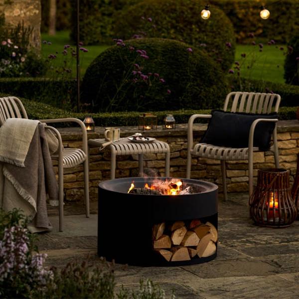 Matte black round steel fire pit with a log store base and glowing fire on a stone patio with garden seating.