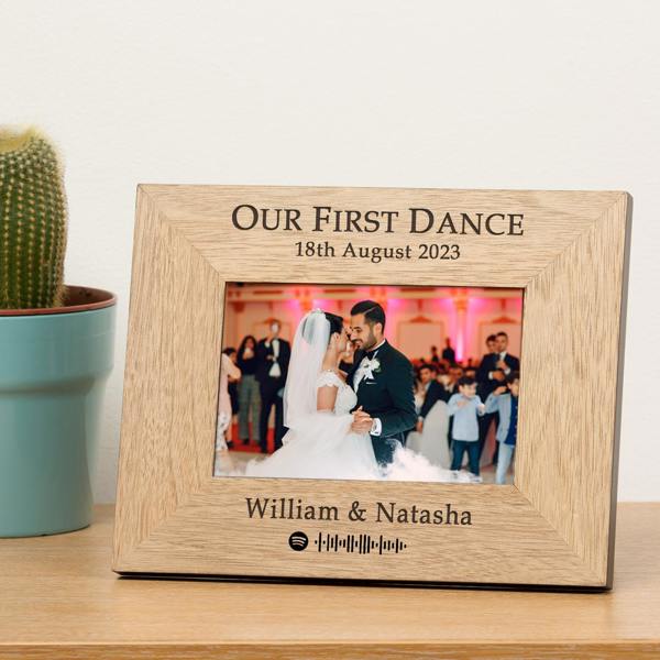A landscape oak veneer wood frame from Abode Home & Gifts engraved with Our First Dance, William and Natasha, and an August 2023 wedding date, displaying a photo of a bride and groom dancing next to a potted cactus on a wooden surface.