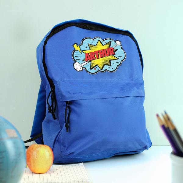 A personalised blue backpack printed with the name ARTHUR, styled on a desk alongside an apple, a notebook, a globe, and coloured pencils.
