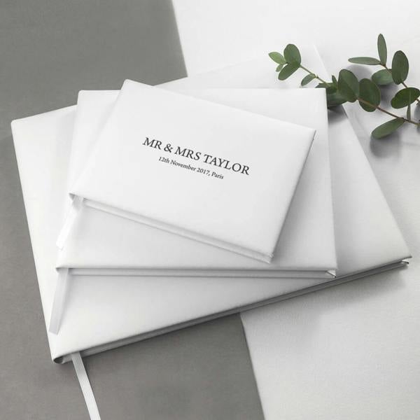 Three sizes of white Italian leather wedding guest books stacked on a two-tone white and grey surface, engraved with Mr & Mrs Taylor, next to a sprig of green eucalyptus.