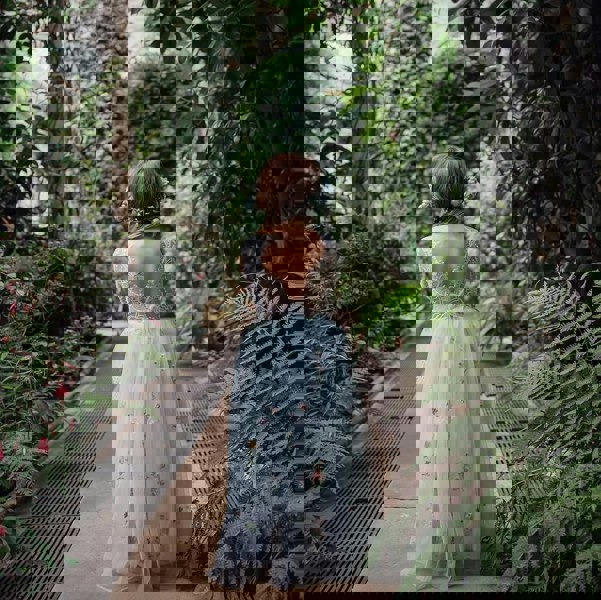 girl in a light blue embroidered dress walking through a lush green garden.