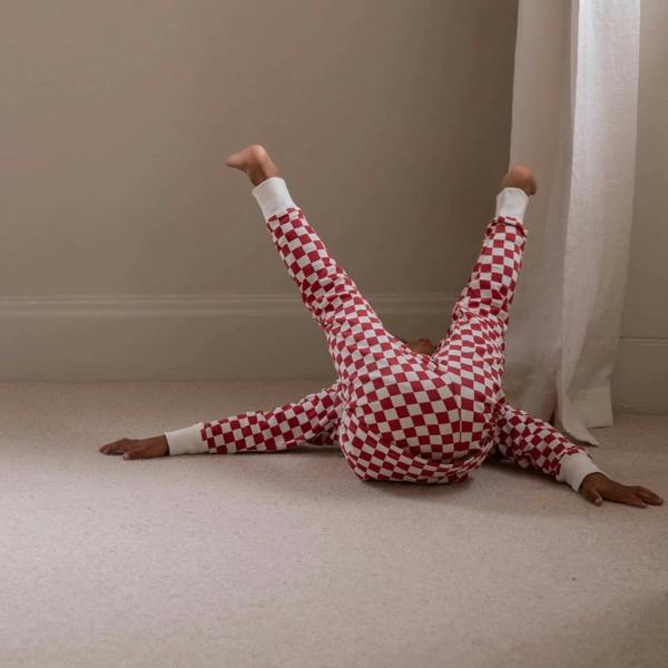 A child lying on their back on a carpeted floor with legs kicked playfully up in the air, wearing matching red and white checkerboard Christmas pyjamas.