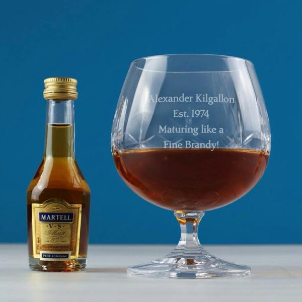 An expertly engraved cut crystal brandy snifter featuring the name "Alexander Kilgallon" and the text "Maturing like a Fine Brandy!" sitting next to a miniature bottle of Martell VS cognac on a light wooden surface against a vibrant blue background.