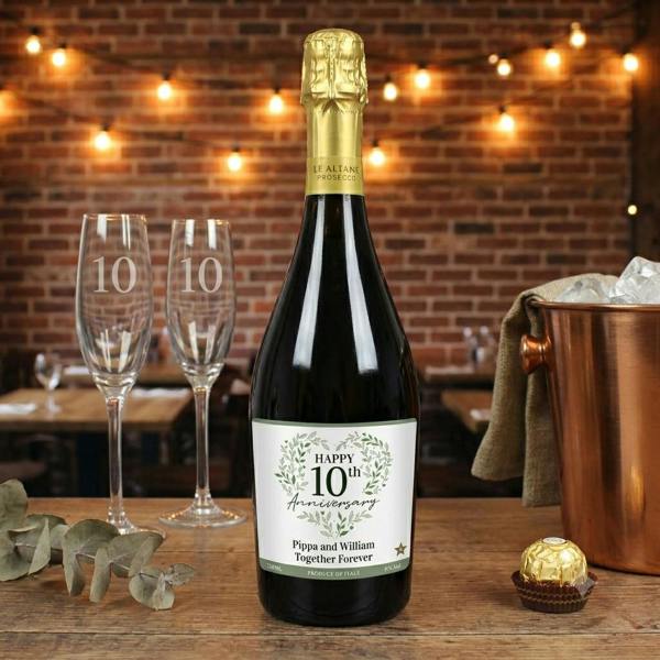 A bottle of personalised Botanical Anniversary Prosecco from Abode Home & Gifts featuring a custom green leaf 10th anniversary label reading Pippa and William Together Forever, displayed on a rustic wooden table next to matching engraved 10 champagne flutes and a gold ice bucket.