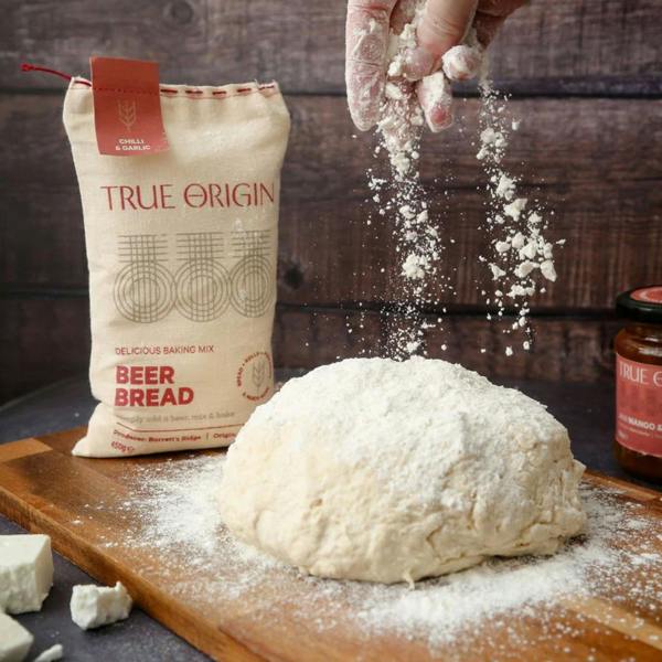 A hand sprinkling flour over a round ball of artisanal beer bread dough on a floured wooden chopping board, next to a fabric sack of baking mix and a jar of chutney.