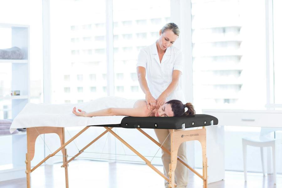 A woman enjoys a massage from a therapist in a bright, modern room, lying face down on the Rio Professional Ultra-Light Portable Massage Table covered with crisp white linens.