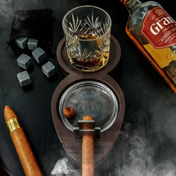 Overhead shot of a dark wooden whisky and cigar tray set with a filled glass tumbler, an ashtray holding a cigar, a cigar cutter, and loose soapstone whisky rocks on a dark table.