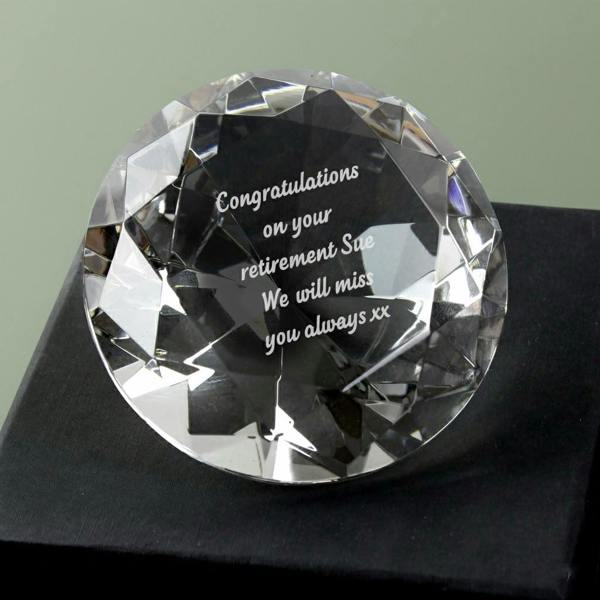 A clear, faceted glass diamond paperweight resting on a black box, featuring white engraved text: "Congratulations on your retirement Sue We will miss you always xx".