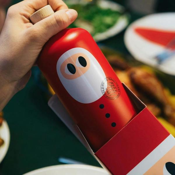 Close-up of a hand lifting the 500ml Santa edition extra virgin olive oil bottle out of its custom Santa gift box at a Christmas dinner table.