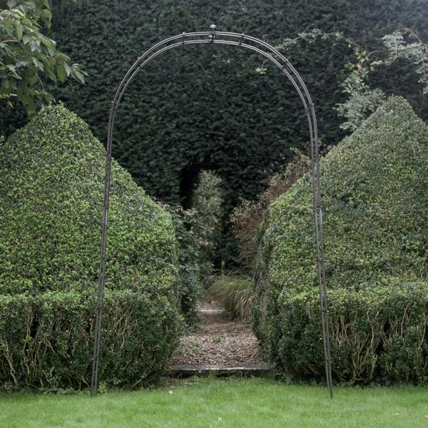 A tall metal rose arch with a curved top and ball finial, positioned between two large green shrubs.