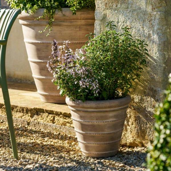 Close-up of the small 36cm Cotswold ribbed planter in washed terracotta ficonstone, planted with green herbs and pale purple flowers on a gravel patio.
