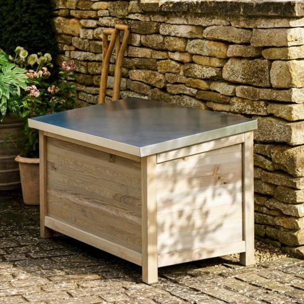 A weathered grey wooden outdoor storage box with a reflective, flat metal lid. The rustic timber cabinet is shown on a wooden deck, positioned next to a black exterior wall with a large garden spade leaning against it. This durable outdoor furniture piece is ideal for organizing garden tools and summer entertaining essentials like picnic blankets and outdoor cushions.