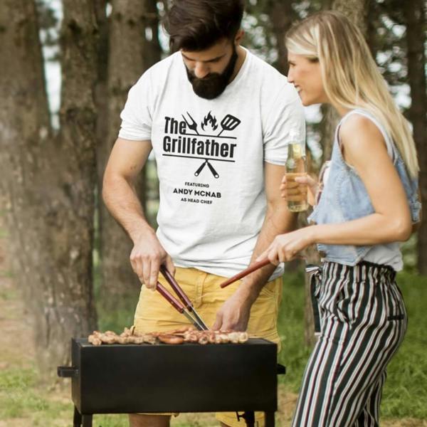 A man with a beard wearing a white personalised 'Grillfather' t-shirt with 'Featuring Andy McNab as Head Chef' printed on the front, standing by an outdoor barbecue with a woman holding a bottle of beer.