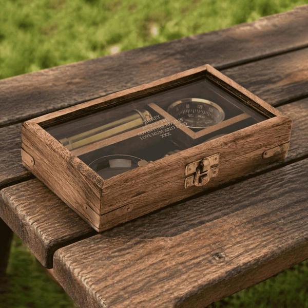 A personalised brass explorer gift set featuring an engraved wooden box with a glass lid displaying a brass telescope, compass, and magnifying glass on a rustic bench.
