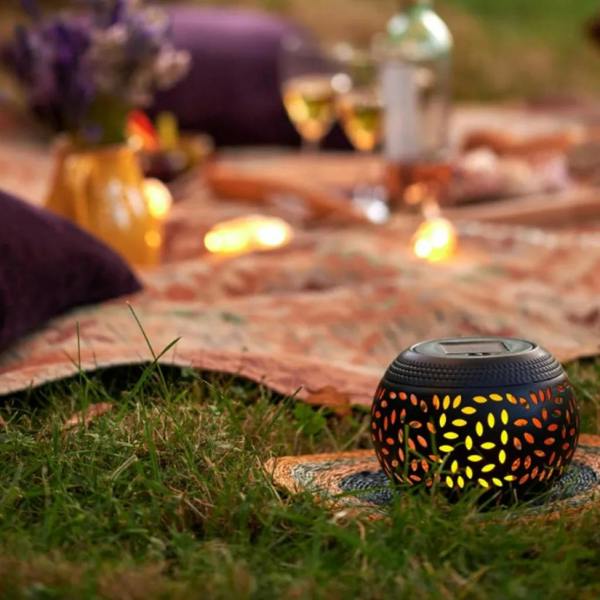 A close-up of a spherical Moroccan solar powered filigree flame lantern glowing with intricate amber light patterns on a dark wooden table.