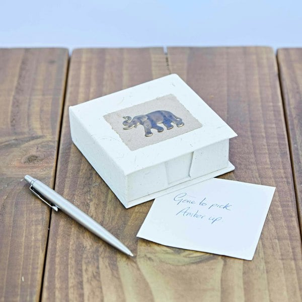 On a wooden surface lies an Elephant Dung Memo Box in white with an elephant image, alongside a silver pen and a note that reads "Gone to pick Amber up.