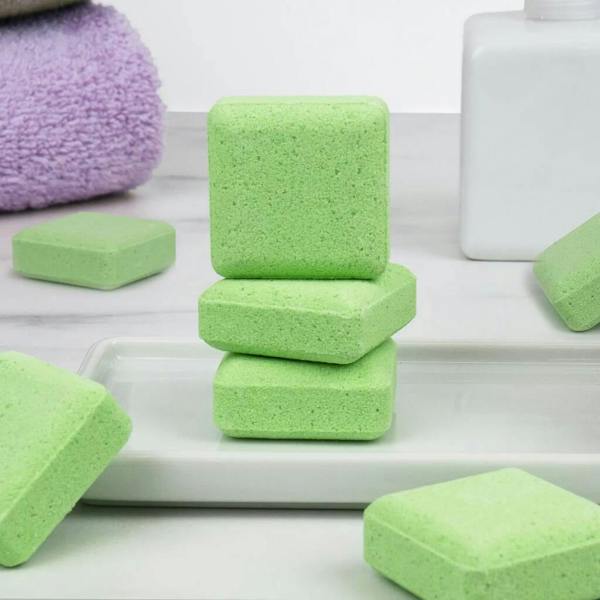 Three square, textured light-green shower steamers stacked neatly on a white ceramic tray, surrounded by more scattered green tablets, a white bottle, and a soft purple towel in the background.
