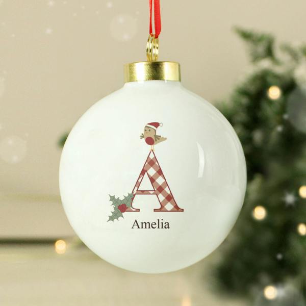 A glossy white ceramic Christmas bauble hanging from a bright red ribbon. The front features a large gingham patterned letter A topped with a cartoon robin in a Santa hat next to holly leaves, with the name Amelia printed below. An out-of-focus Christmas tree with warm lights fades into the background.