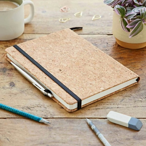A personalised Natural Cork A5 Notebook on a wooden table with pens, pencils and a cup of coffee.