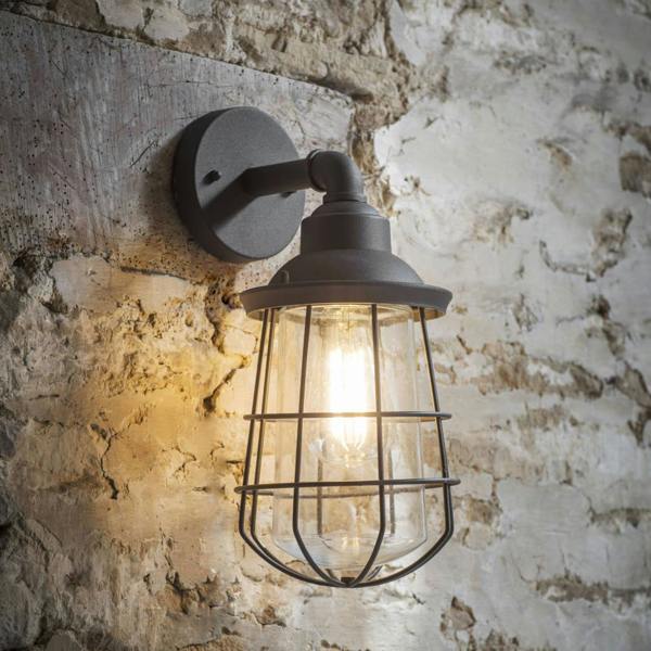 An industrial matte charcoal outdoor cage wall light illuminated with a warm filament bulb against a rustic brick wall.