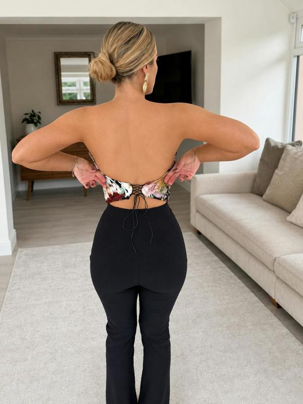 A woman with blonde hair in a bun adjusts the tie of her Sarvin Ivy Corset Top, featuring a waist-cinching floral print. She pairs it with black high-waisted pants in a bright room with neutral decor, standing with her back to the camera.