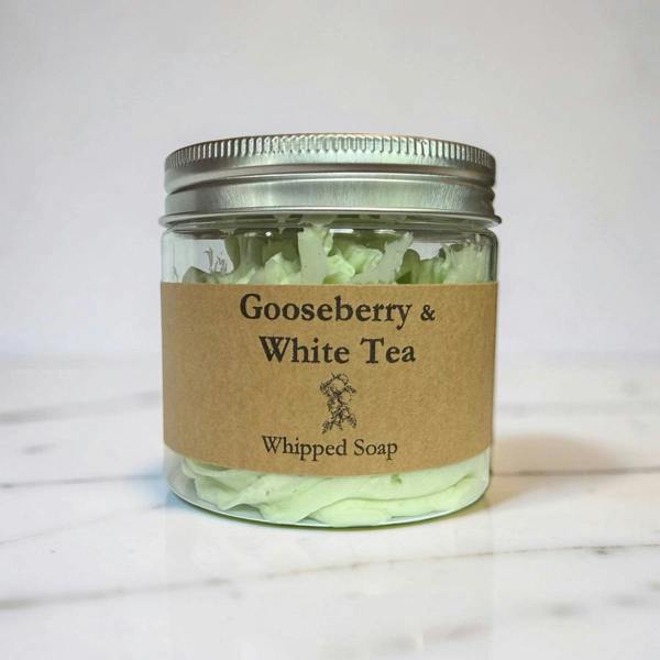 A clear glass jar of Gooseberry & White Tea Whipped Soap with a brown kraft paper label and a silver metal lid, set on a white marble surface against a white background.