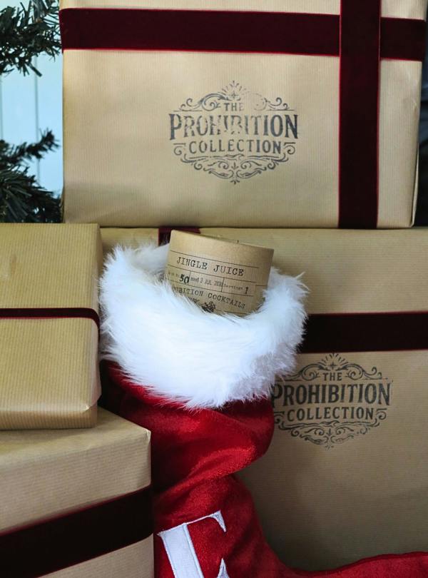 A red Santa-style stocking with white faux fur trim sits among wrapped gift boxes. The stocking contains a small paper ticket reading 'Jingle Juice' by Prohibition Cocktails