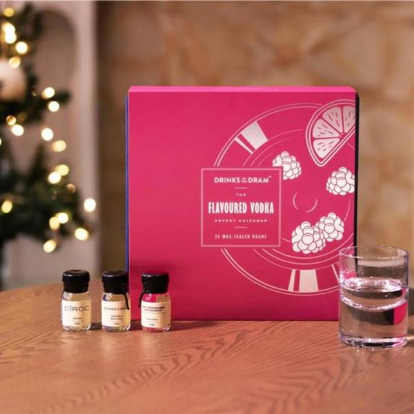 Abode Home & Gifts' Flavoured Vodka Advent Calendar, featuring a bright pink box with raspberry and citrus illustrations, displayed next to three miniature vodka drams and a glass of ice, with a blurred Christmas tree in the background.