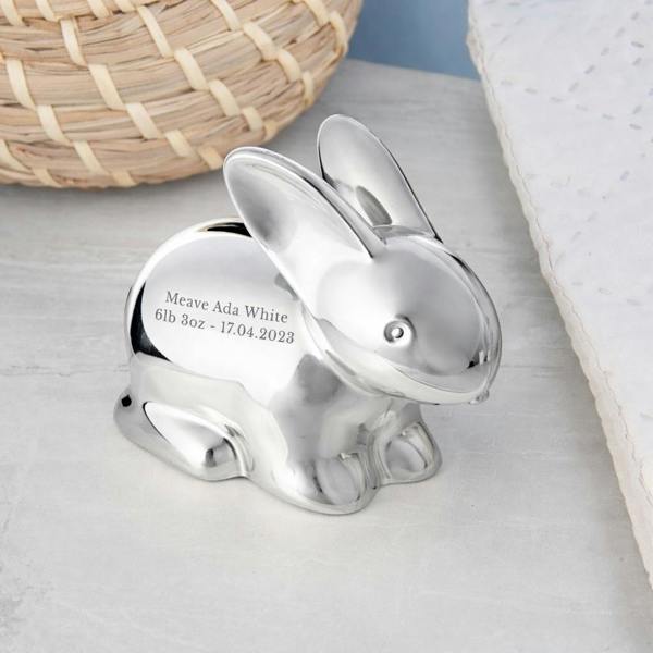 An engraved silver-plated bunny money bank showcasing custom birth details including name, weight, and date, styled on a grey surface next to a woven basket.