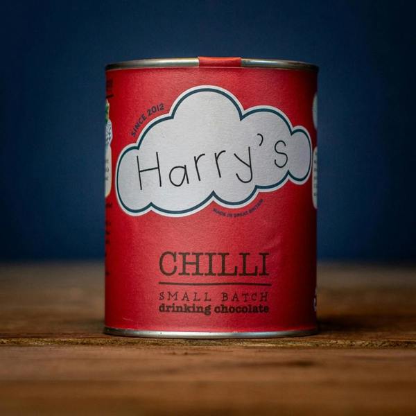 A vibrant red tin of small batch chilli drinking hot chocolate sitting on a rustic wooden table against a dark blue background.